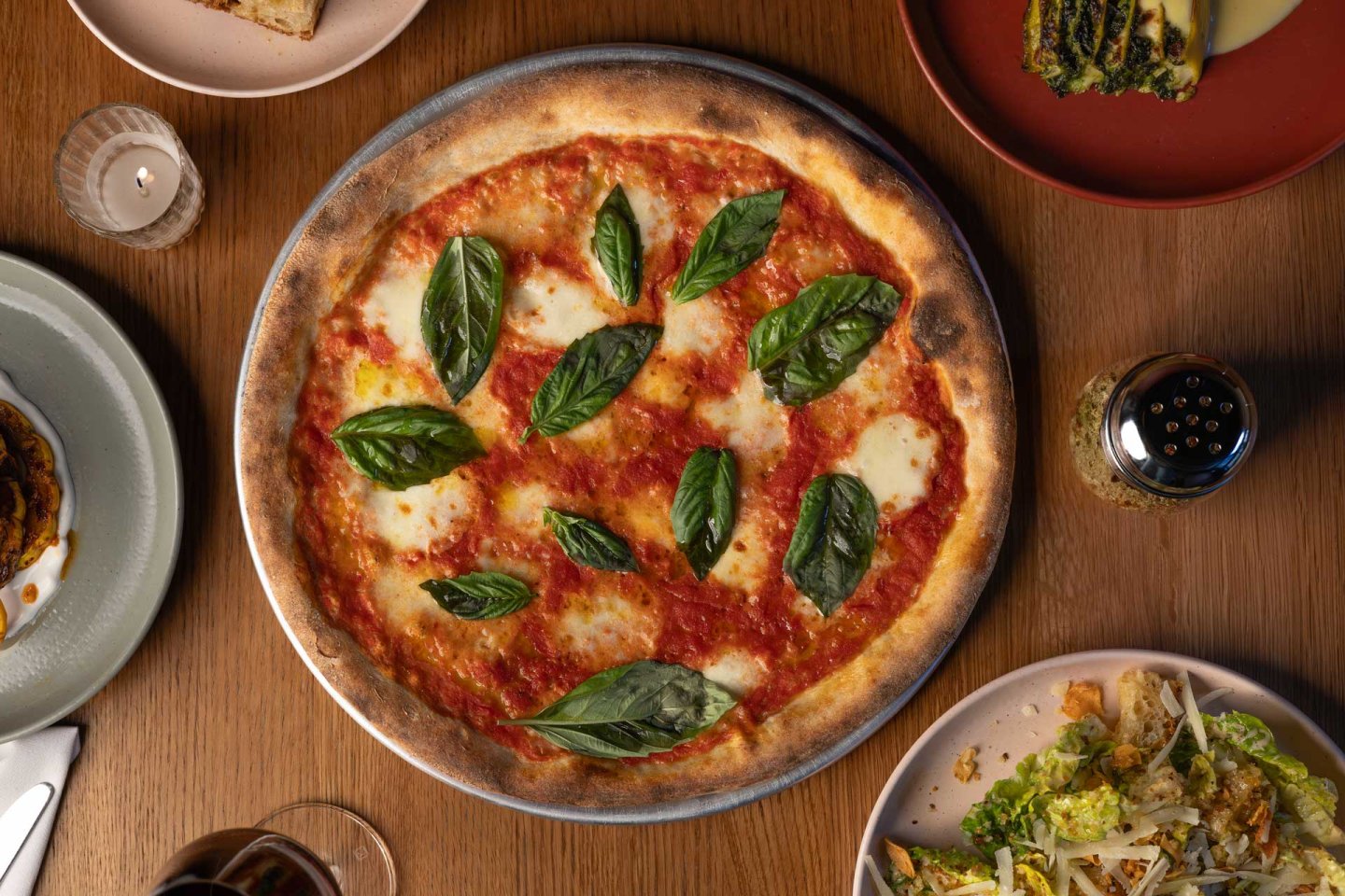 A freshly baked pizza topped with tomato sauce, melted mozzarella, and basil leaves on a wooden table. Surrounding the pizza are plates with food, a lit candle, a pepper shaker, and a glass of red wine.