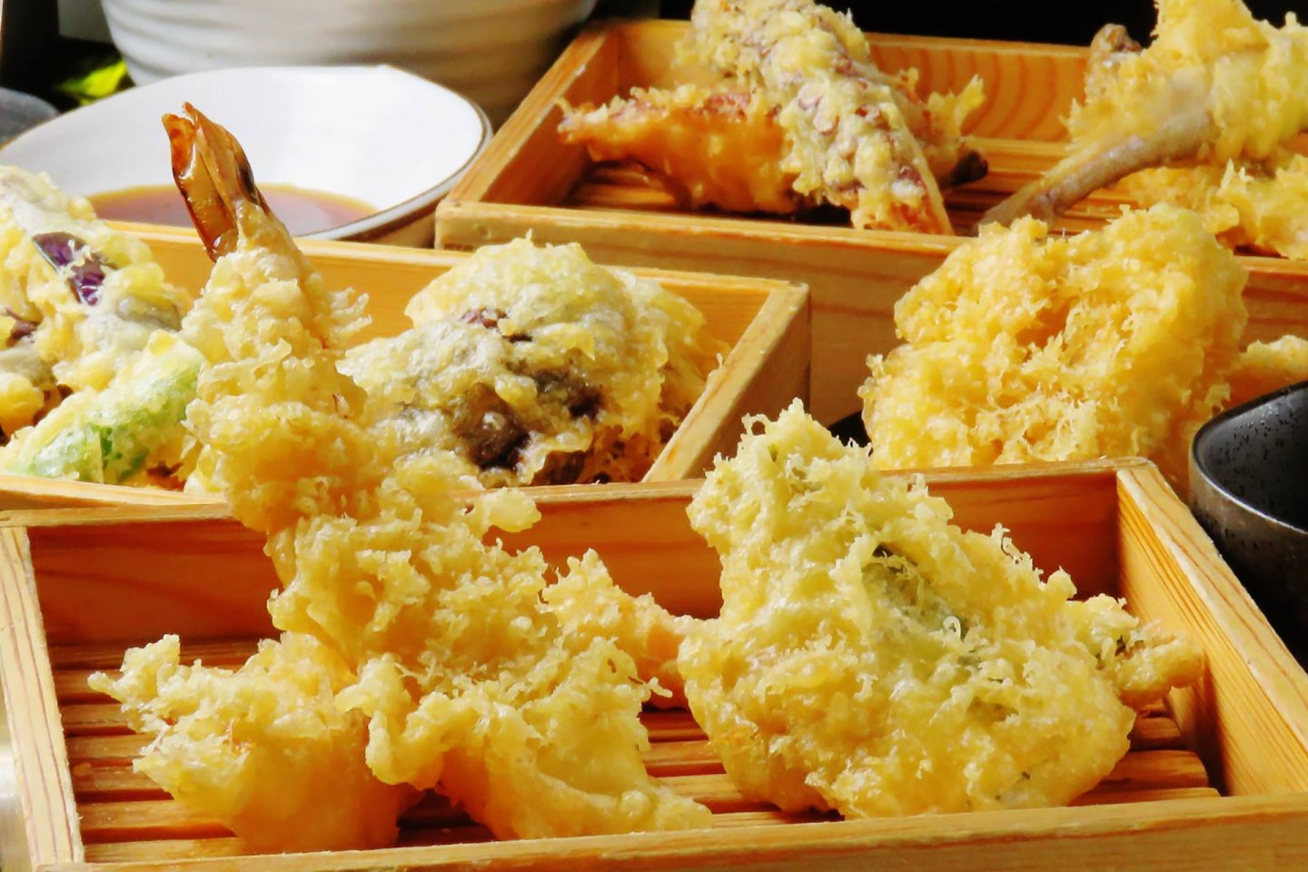 Assorted pieces of golden, crispy tempura including shrimp and vegetables are served in wooden trays, ready to eat, with a bowl of dipping sauce in the background.