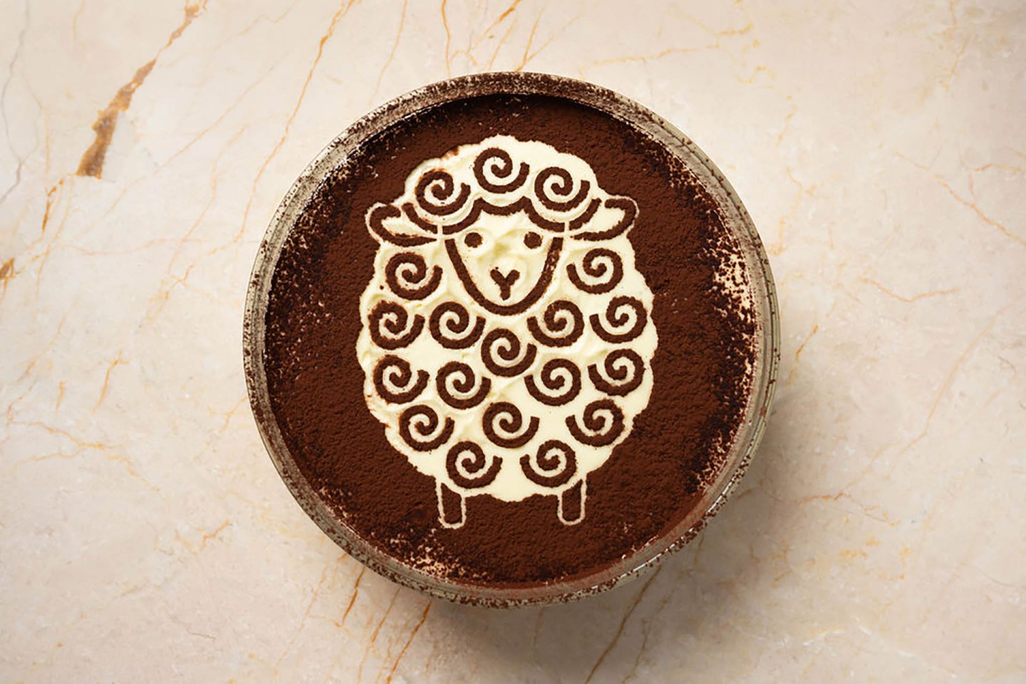 A bowl of dessert dusted with cocoa powder, featuring a cute sheep design made with cream and swirling patterns on top, set on a light marble surface.
