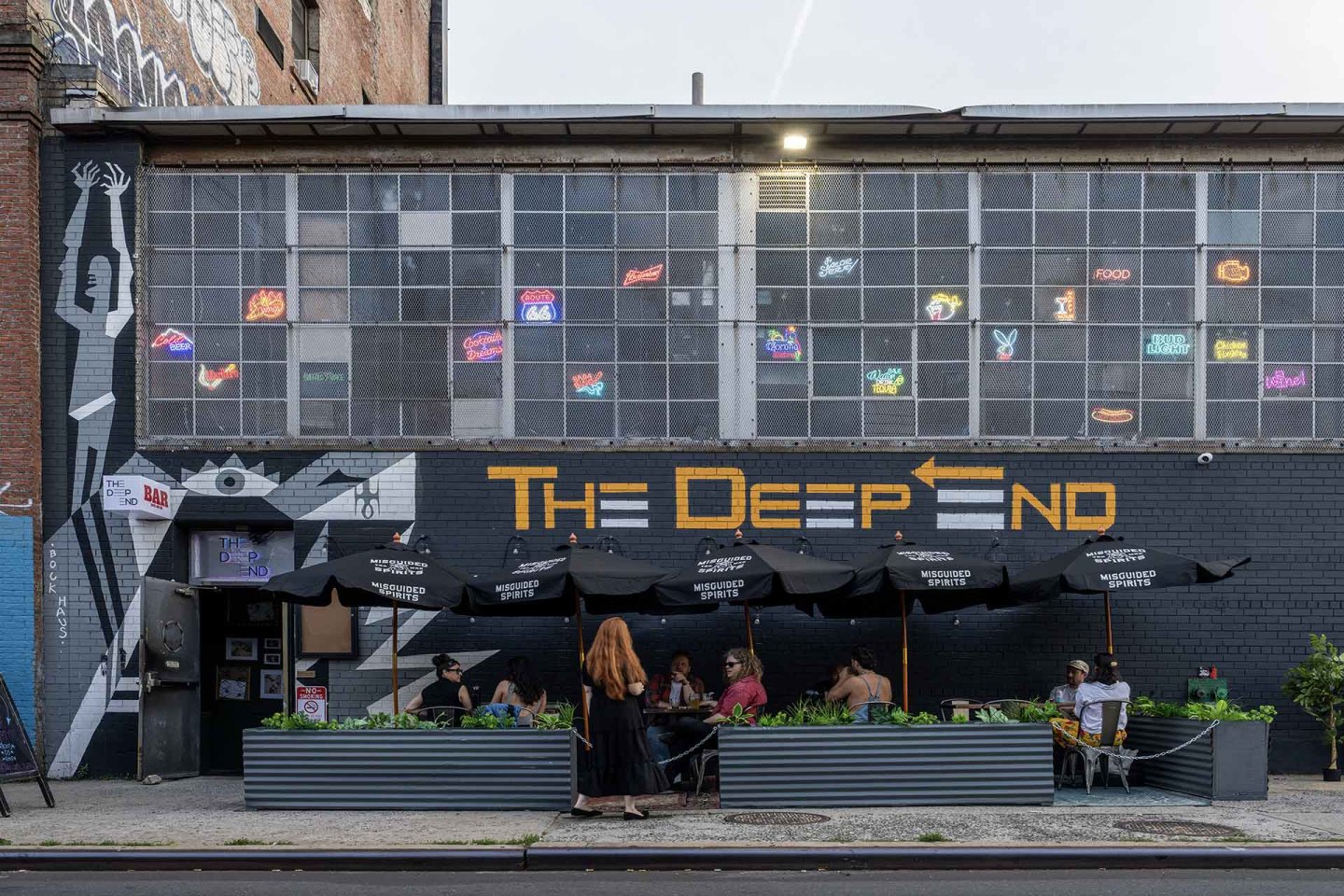 People sit under black umbrellas at outdoor tables in front of “The Deep End” bar, with a mural and neon signs on the building’s upper windows.