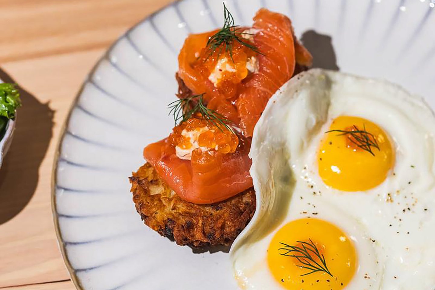 Two sunny-side-up eggs garnished with dill sit beside a crispy hash brown topped with smoked salmon, crème fraîche, salmon roe, and more dill, all arranged on a white plate with a striped rim.