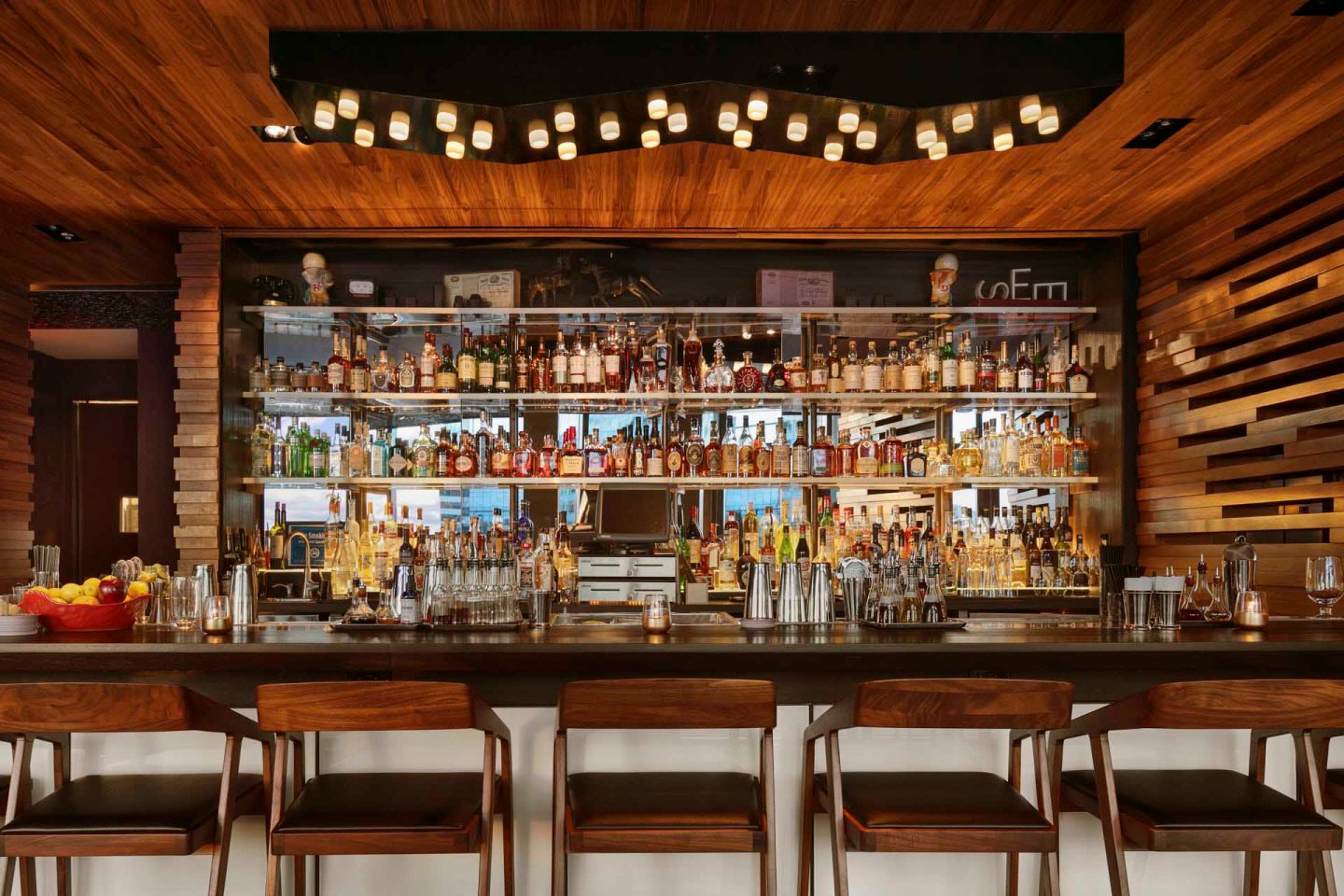 A modern bar with stylish wooden decor featuring a variety of liquor bottles on shelves behind the counter. Three wooden bar stools are in front, and a unique ceiling light fixture adds ambiance
