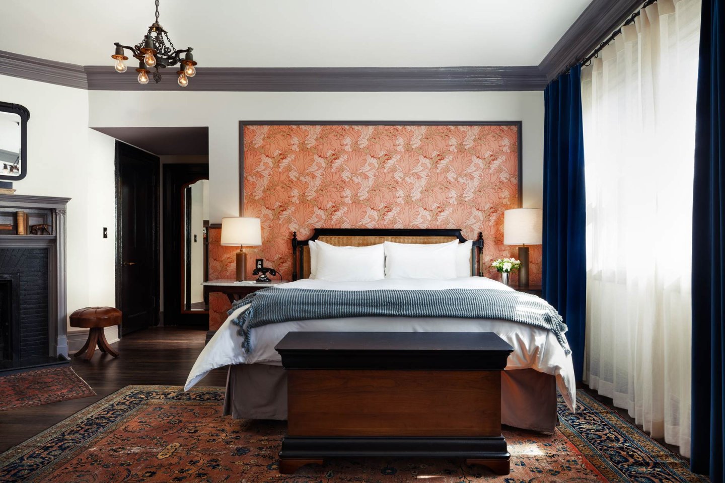 Elegant bedroom with a large bed centered against a pink floral accent wall, flanked by two bedside tables with lamps. A dark bench sits at the foot of the bed, with a large rug and tall windows with blue curtains.