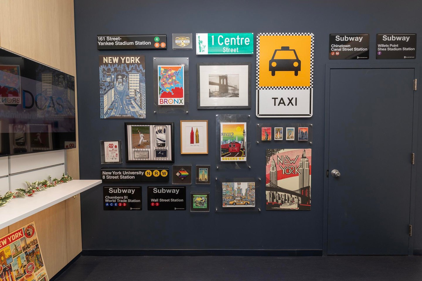 A wall displays framed New York-themed artwork, subway signs, a Bronx poster, images of taxis, and other city memorabilia arranged in a grid pattern beside a closed gray door.