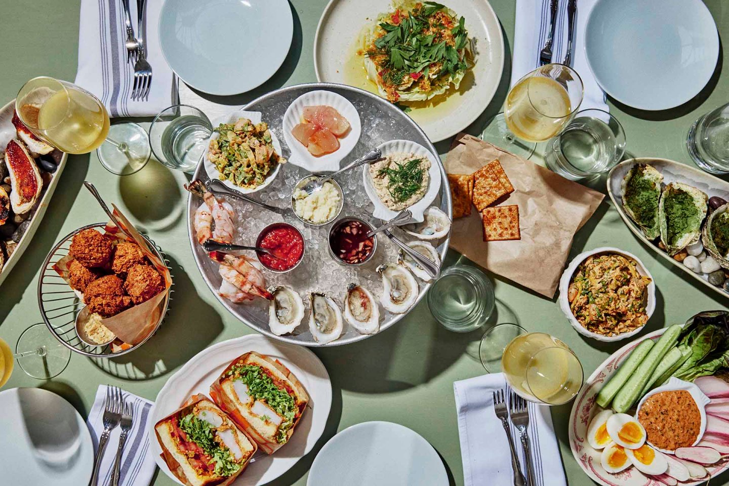A table set with oysters, shrimp, spreads, crackers, sandwiches, falafel, salad, and vegetables, surrounded by plates, napkins, and glasses of white wine.