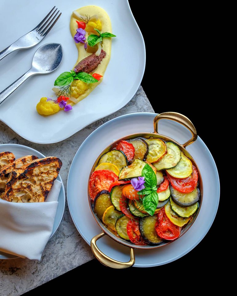 A dish of ratatouille with sliced tomatoes, zucchini, and eggplant, garnished with basil and edible flowers, is served alongside grilled bread and an elegant plated appetizer with colorful garnishes.