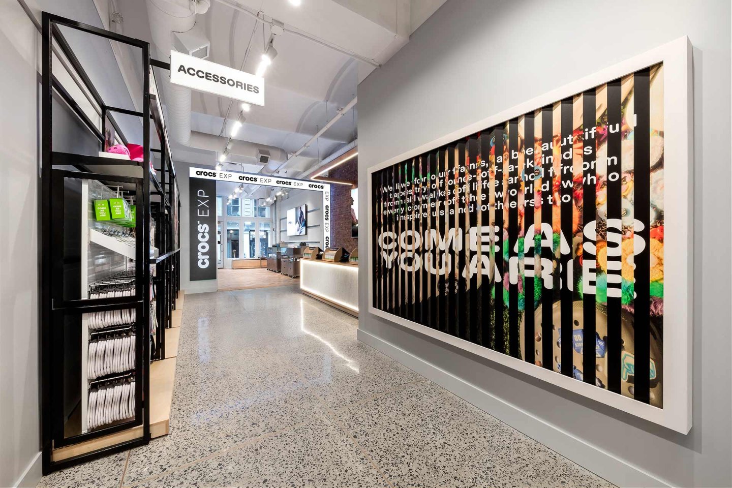 Modern retail store with high ceilings, bright lighting, a large “Accessories” sign, organized shelves, and a colorful Crocs display wall featuring a bold, partially obscured slogan. Reception counter visible in the background.