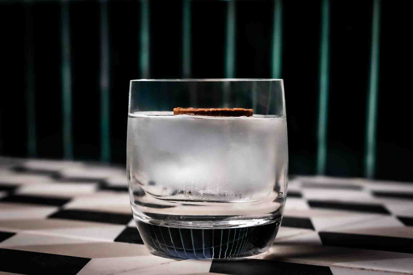 A clear glass with ice water and a round slice of dried citrus sits on a black-and-white patterned surface, with vertical green lines in the blurred background.