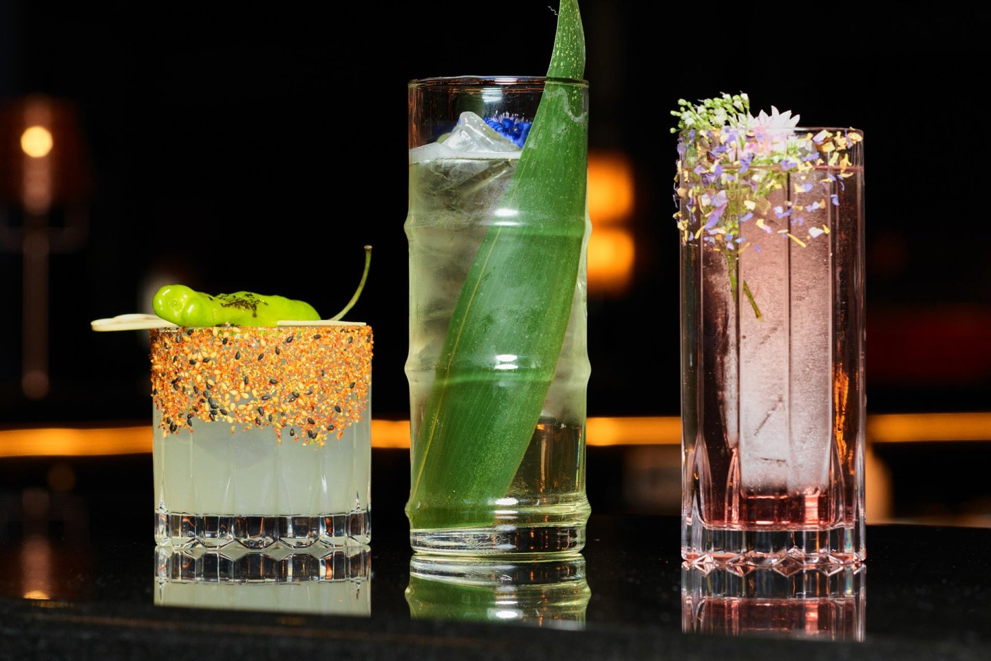 Three cocktails are displayed: a short glass with a spicy rim and green pepper garnish, a tall glass with a leaf and blue flower, and another tall glass with pink liquid and floral garnish, all on a reflective surface.