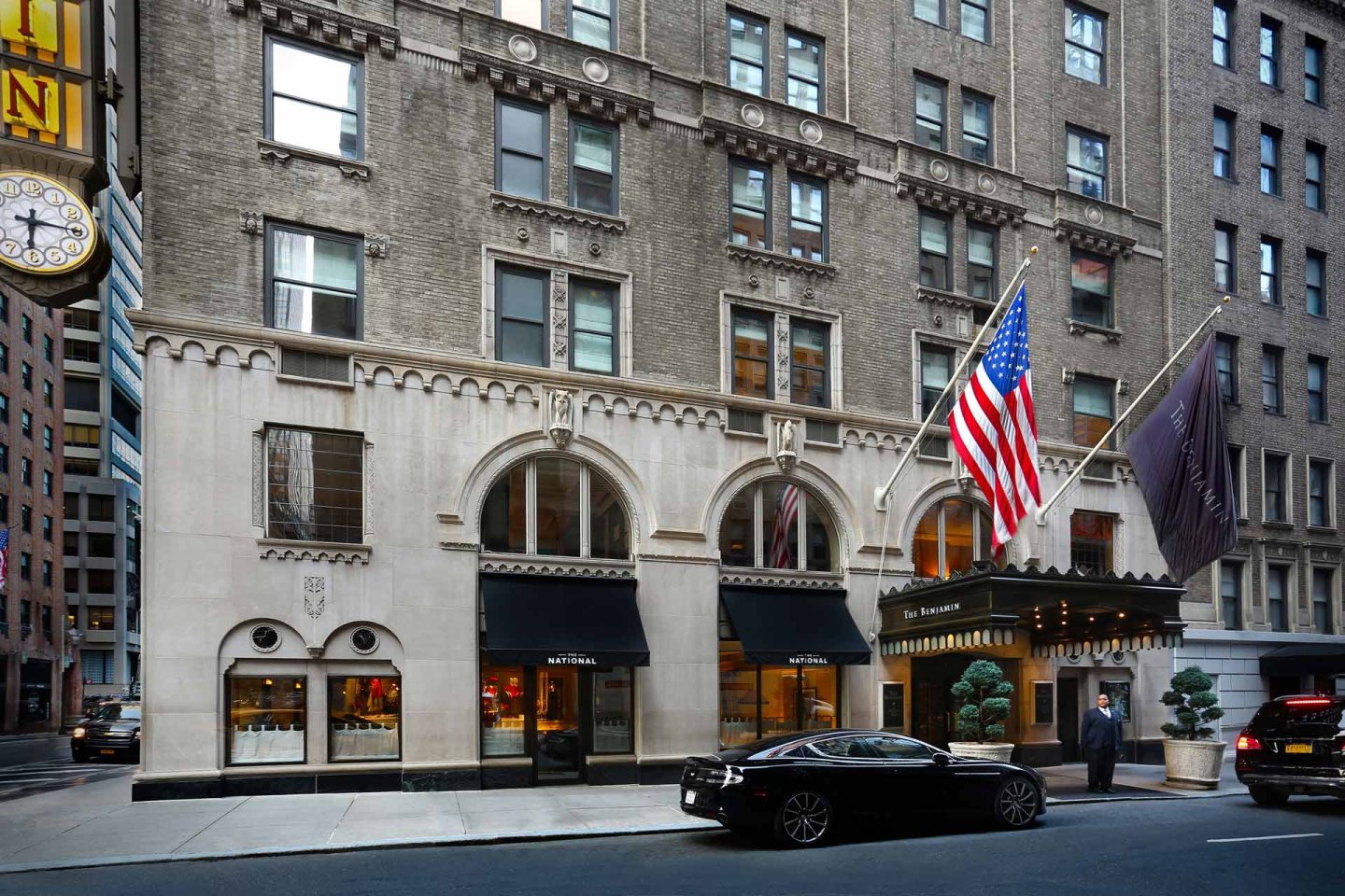 Street view of a historic hotel with tan brick facade, large arched windows, two flags (American and hotel), a valet standing by the entrance, and a black car parked in front.