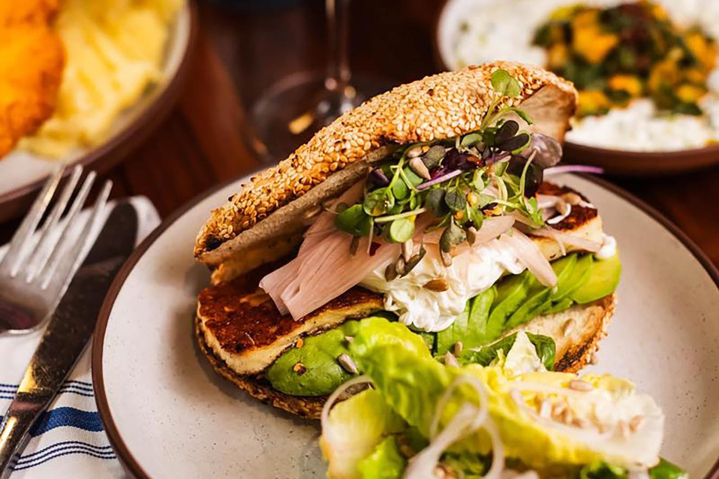 A close-up of a sesame seed sandwich bun filled with leafy greens, halloumi cheese, microgreens, and pickled onions, served with a fresh green salad on a plate.
