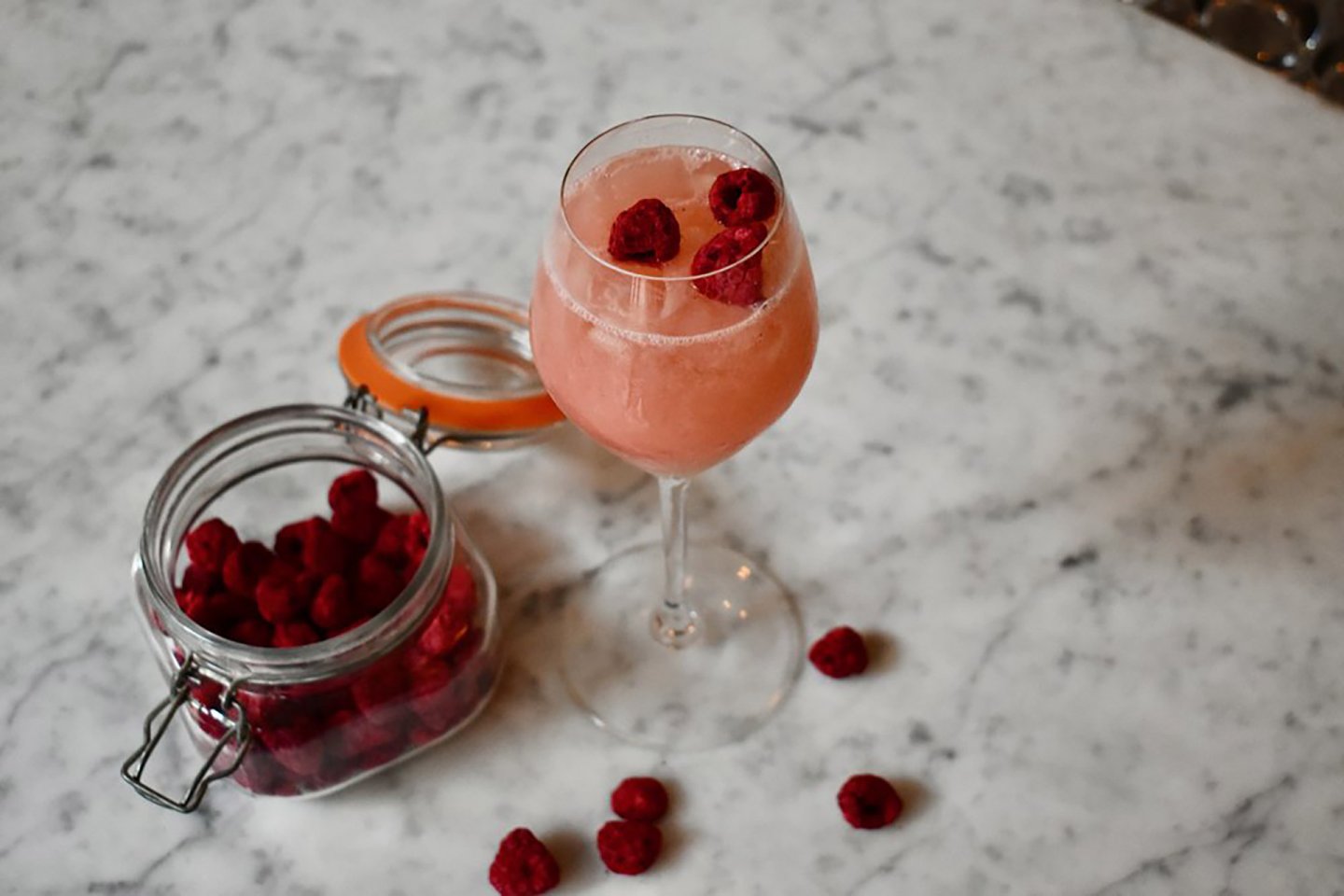 A glass of pink drink with ice and raspberries sits on a marble surface next to an open jar of raspberries, with a few raspberries scattered around.