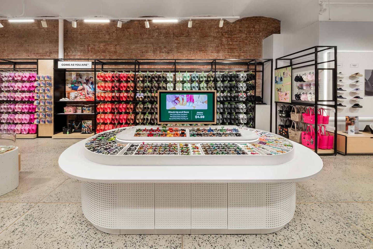 A brightly lit retail store with colorful footwear and bags displayed on wall racks, a central white display table filled with accessories, and a digital screen showcasing promotions. Brick and white walls create a modern aesthetic.