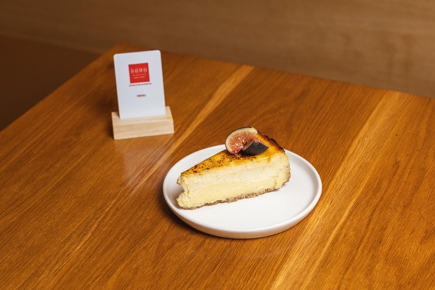 A slice of cheesecake topped with a fig slice sits on a white plate on a wooden table. A small card in a wooden holder is in the background.