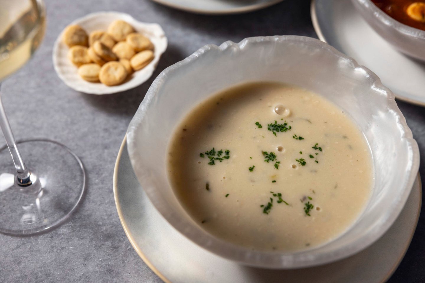 A bowl of creamy soup garnished with chopped herbs is served on a gray table. Nearby are a glass of white wine and a small dish of round crackers. The setting creates an elegant dining ambiance.