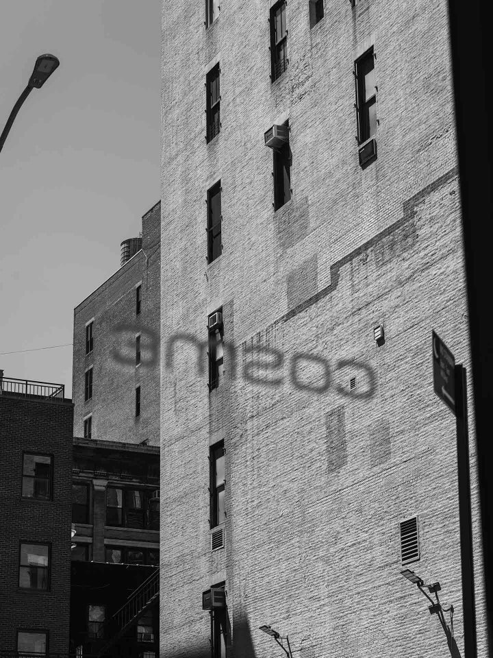 A black and white photo of tall brick buildings. The shadow of a sign reading "Cosine" is cast on one of the buildings. A streetlamp and portions of fire escapes are also visible.