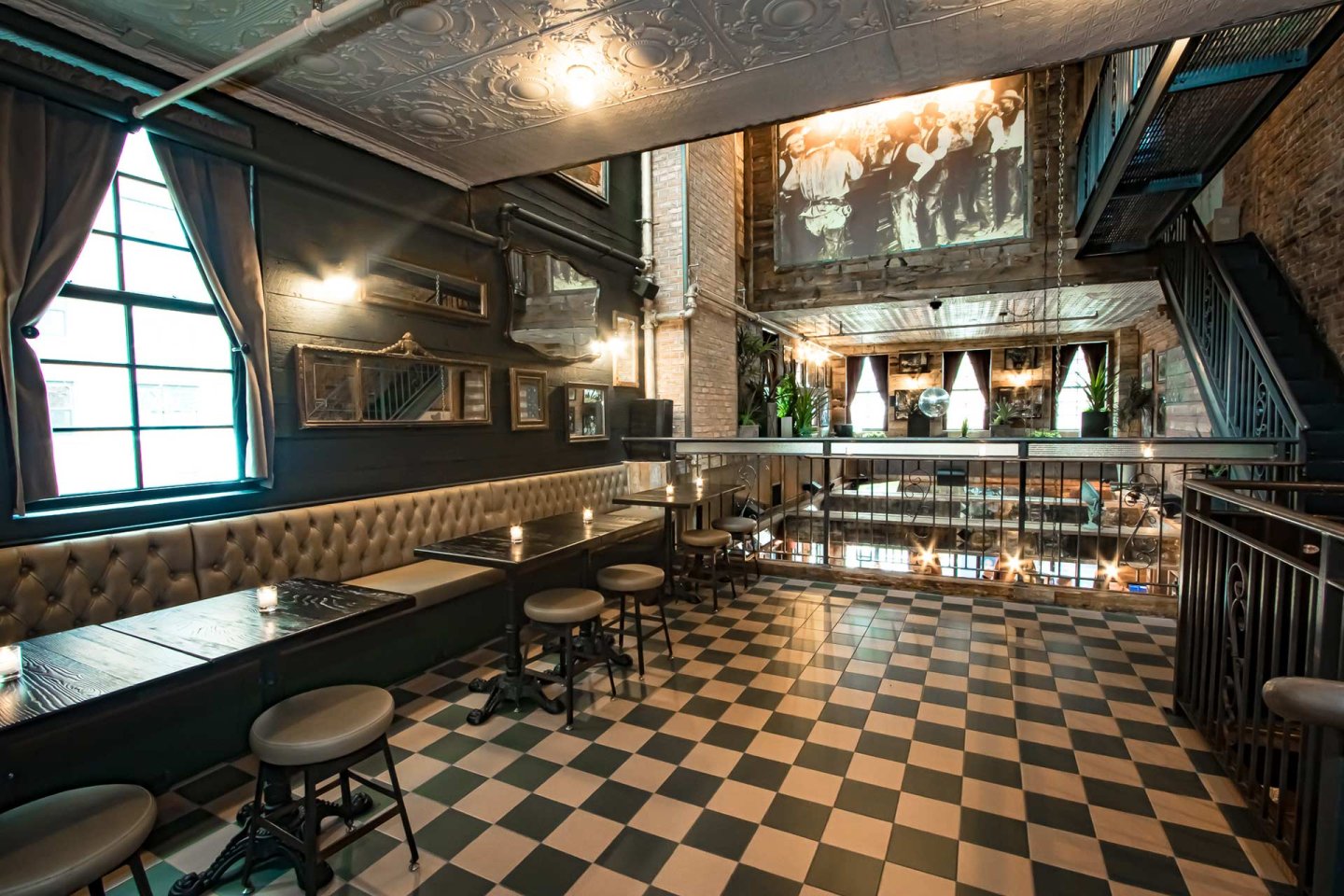 A stylish, vintage-inspired bar with checkered floors, tufted bench seating, and small round tables. Large mirrors, framed photos, and a sepia-toned mural decorate the walls. Natural light streams in from large windows.