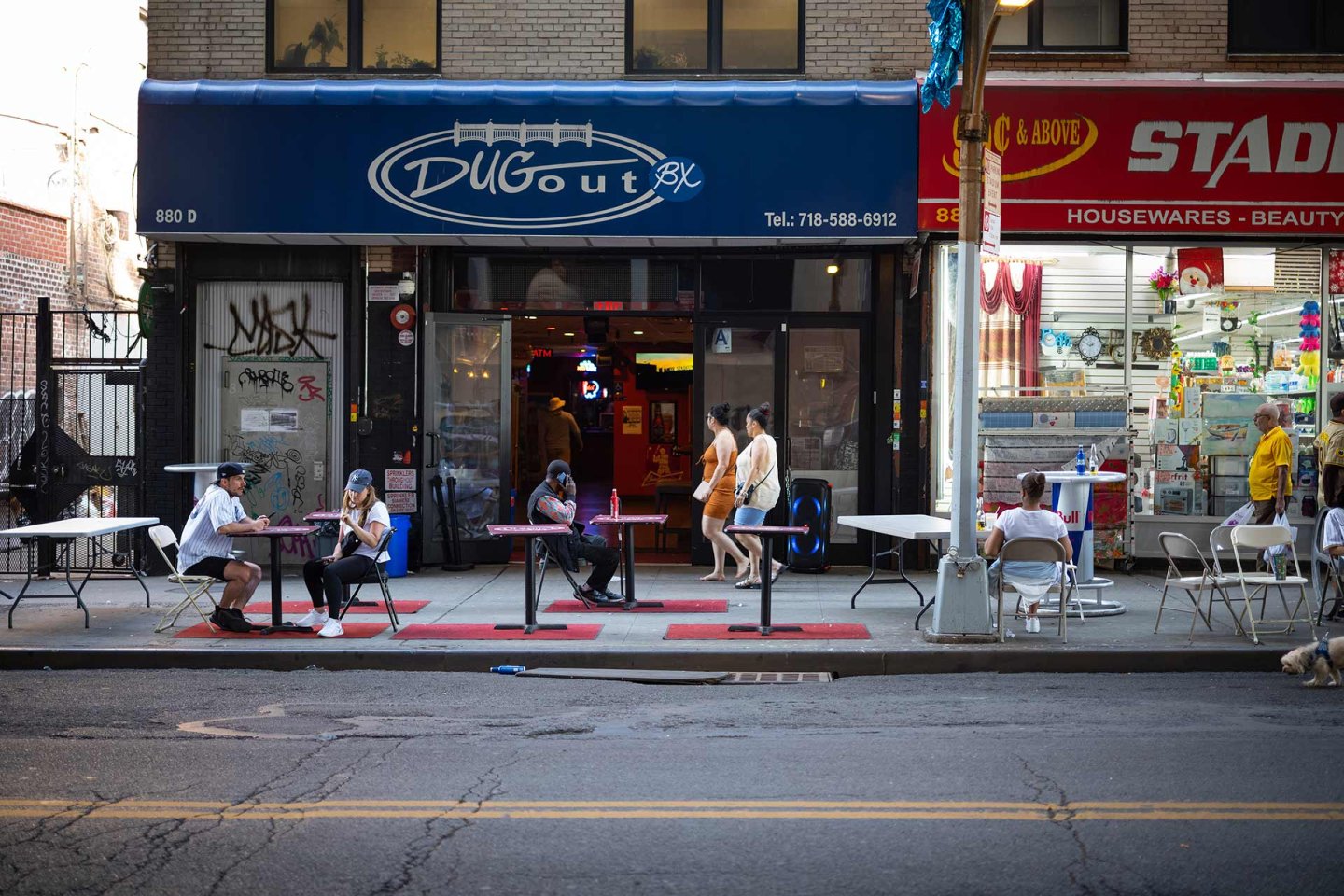 People sit at tables on a sidewalk outside Dugout bar; others walk past. The bar has a blue awning, and a brightly lit store selling housewares is next door. The street is quiet with little traffic.