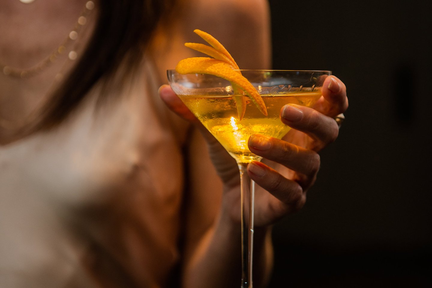 A person in a sleeveless top holds a cocktail glass filled with a yellow drink, garnished with a twist of orange peel. The background is dark and out of focus.