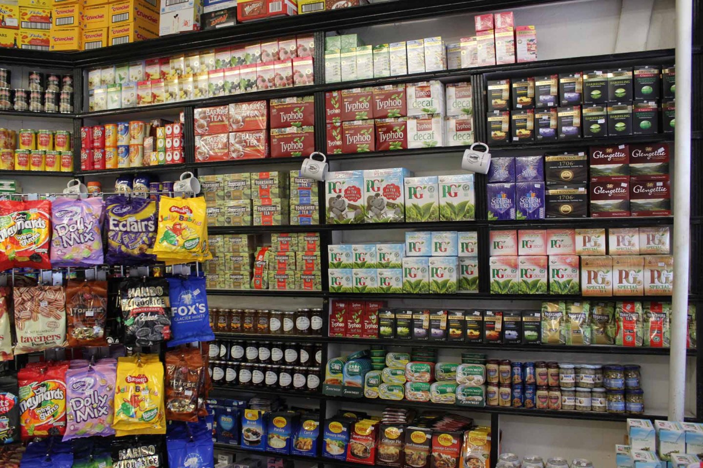 Shelves in a store stocked with various brands of tea, coffee, biscuits, and other packaged snacks. Assorted colorful packets and boxes are neatly arranged, with tea and coffee products prominently displayed.