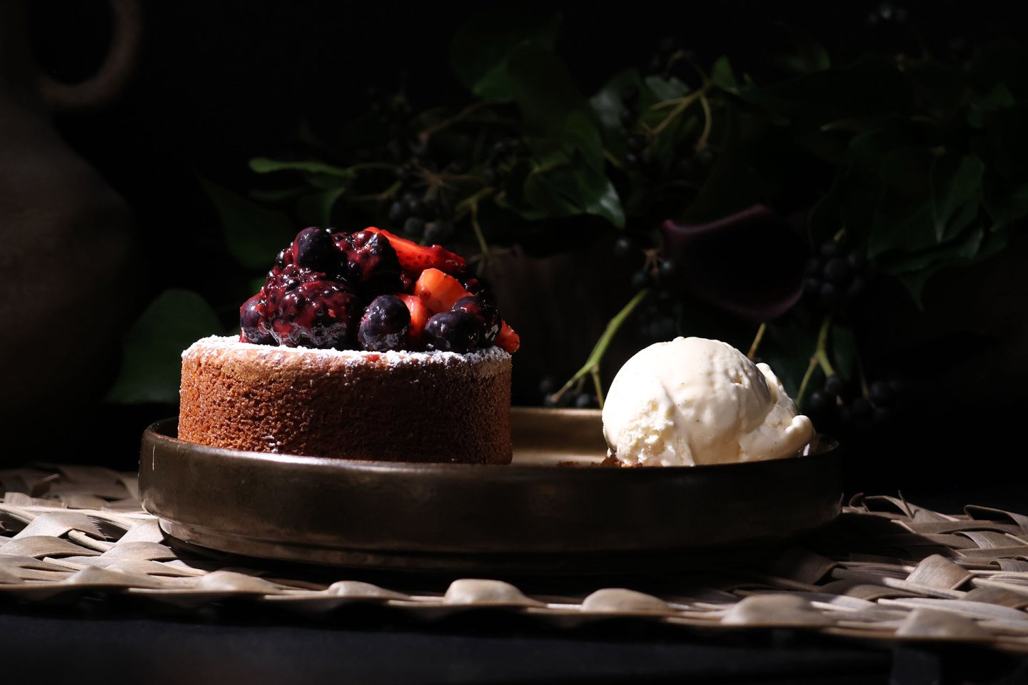 A small cake topped with mixed berries sits on a round plate next to a scoop of vanilla ice cream, with leaves and dark background.