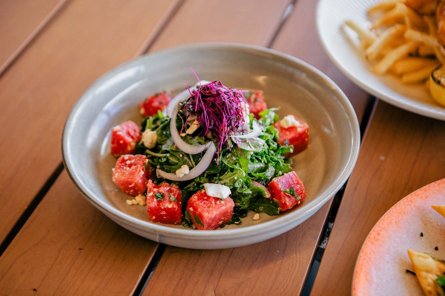 A bowl of salad with watermelon cubes, mixed greens, sliced onions, crumbled cheese, and a garnish of purple sprouts, served on a wooden table.