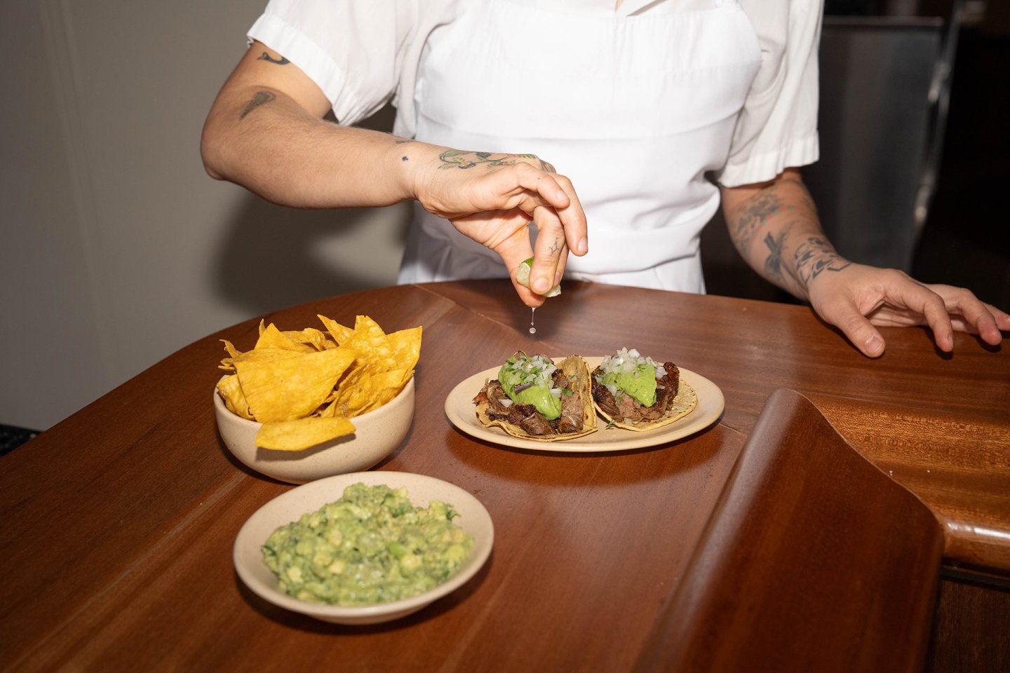 A person in a white apron squeezes lime over two tacos on a plate. Nearby, there are bowls of tortilla chips and guacamole on a wooden counter.