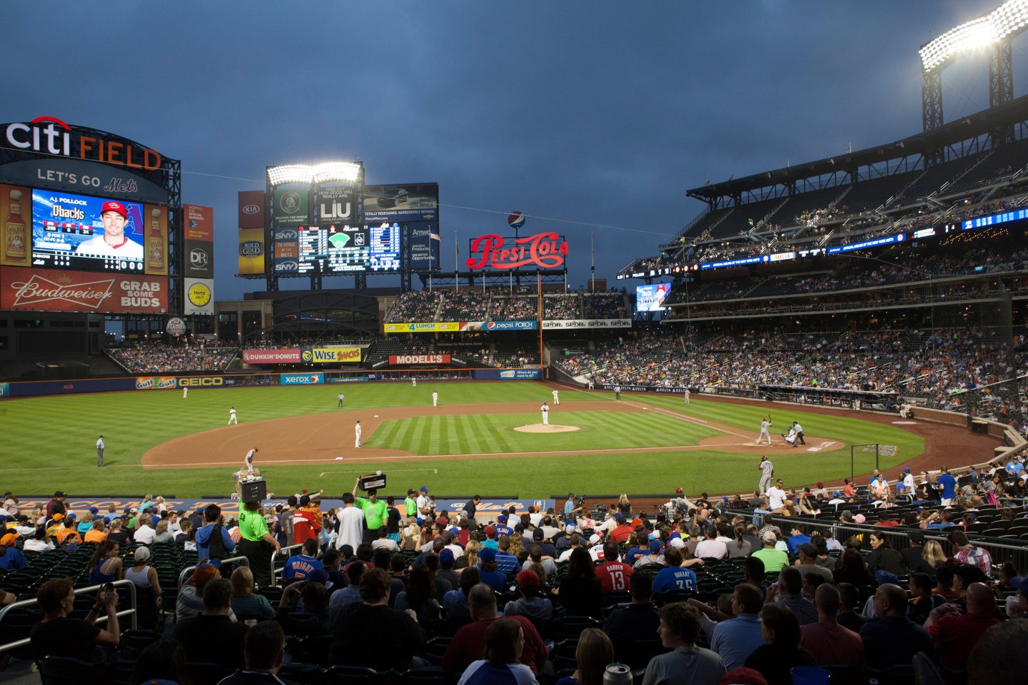 A lively baseball game at a stadium during dusk. The field is illuminated under darkening skies, with large screens displaying advertisements. Spectators fill the stands, watching the action on the field.