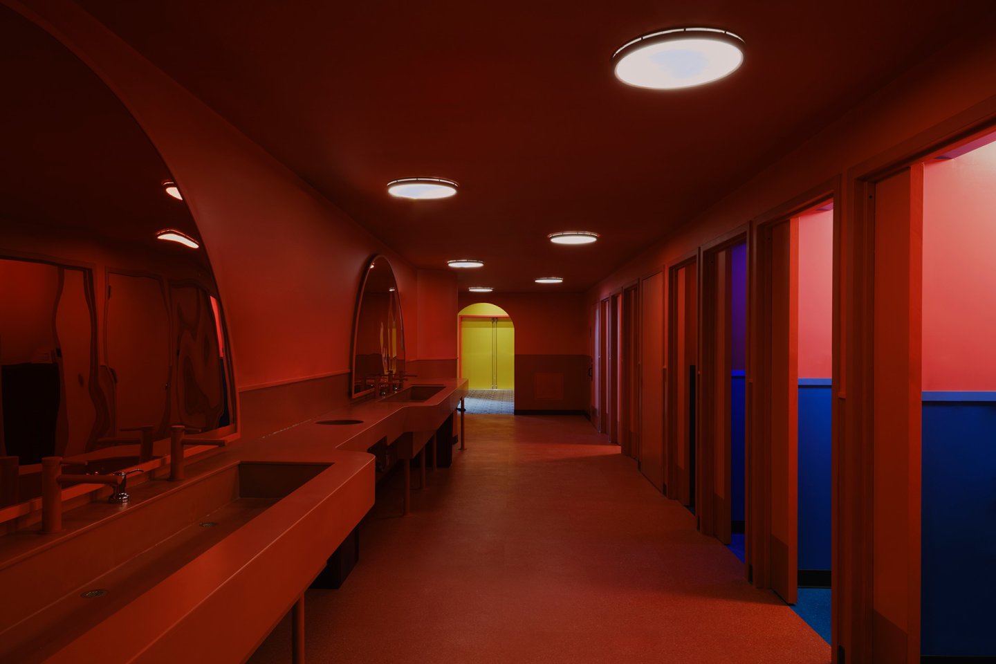 A brightly lit, modern restroom with red walls and sinks, circular ceiling lights, large mirrors, and partially open stall doors revealing blue interiors. Yellow double doors are visible at the end of the hallway.