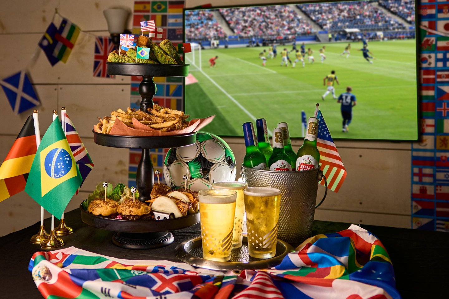 A three-tier tower of shareable matchday bites at BLACKBARN: crispy vegetable samosas, parmesan fries, sliders, bao buns, shrimp lettuce wraps with a bucket of ice-cold beers, a soccer ball and national flags, with a soccer match playing on a screen behind.