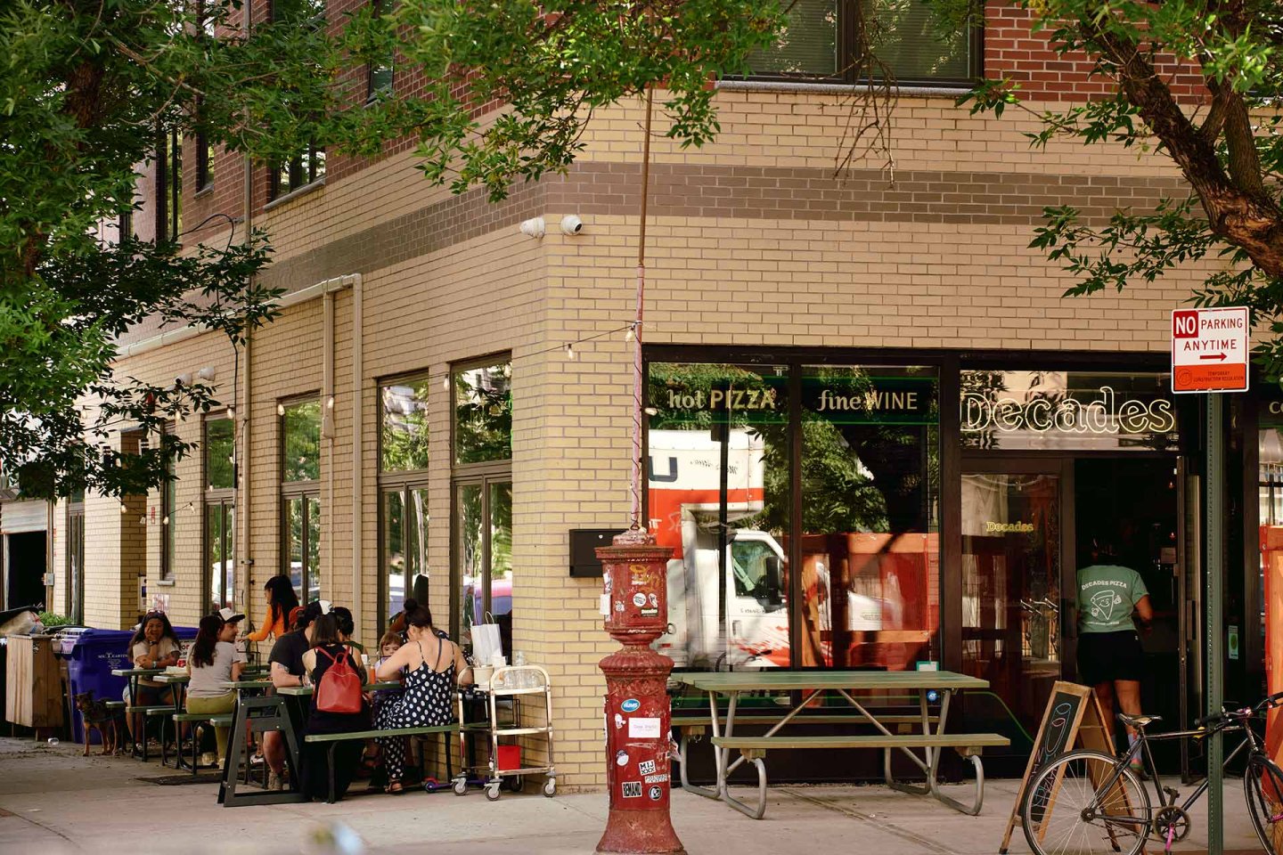 People dine outside a pizza and wine restaurant called “Decades” on a city sidewalk. Trees provide shade, and tables are set up along the building. A “No Parking Anytime” sign and a red fire call box are visible.