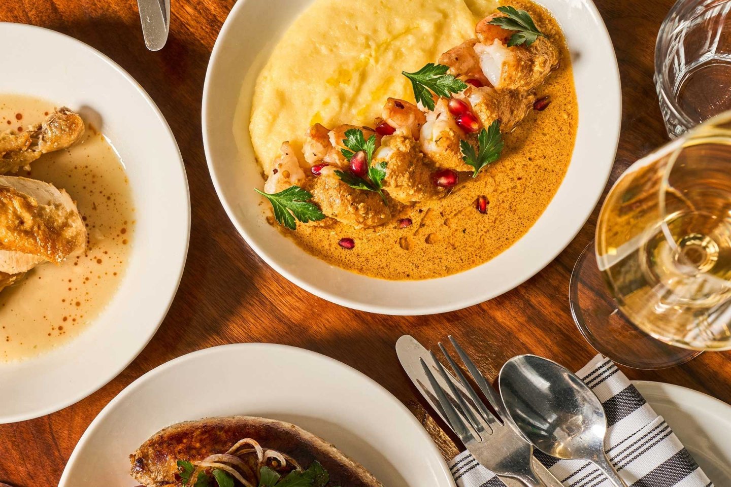 A table set with plates of gourmet food, including a creamy polenta dish topped with shrimp, sauce, and garnishes, alongside other plated dishes, a glass of white wine, and silverware on a napkin.