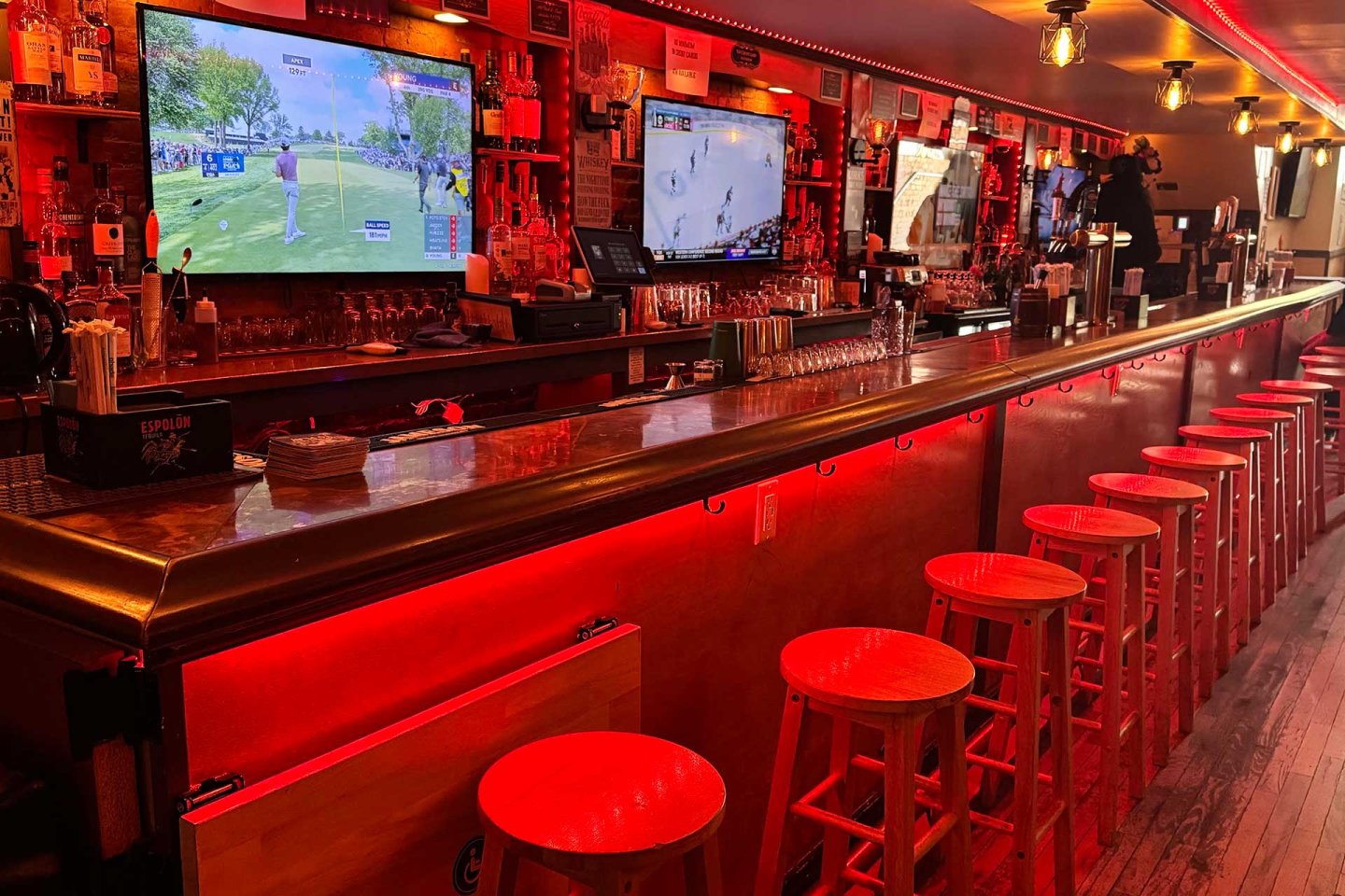 A long bar with red lighting, wooden stools lined up, and two large TVs showing sports above the counter. Glasses, bottles, and bar equipment are visible on the shelves behind the bar.