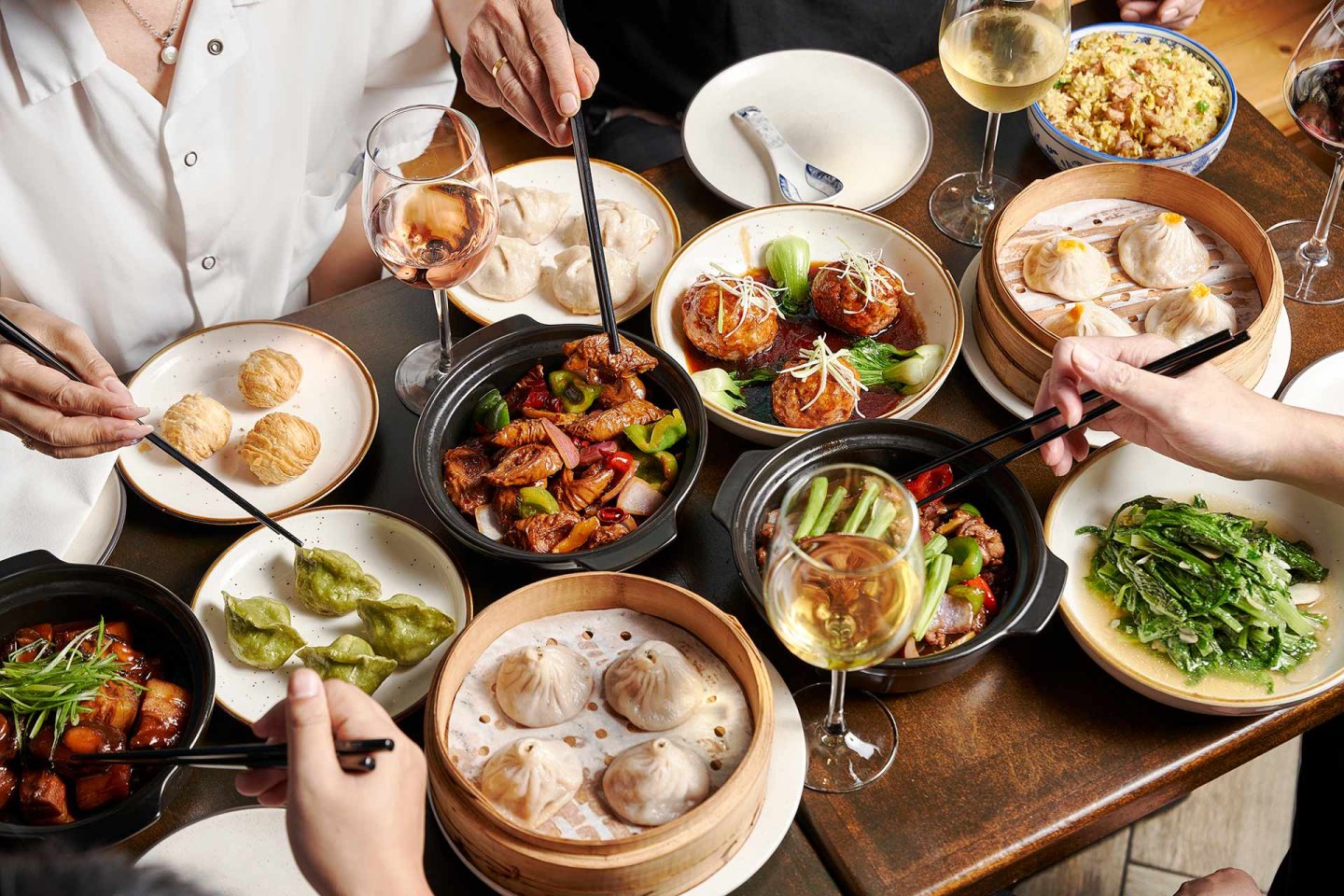 A group of people share a variety of Chinese dishes, including dumplings, stir-fried vegetables, meat dishes, and glasses of white wine, all served on a wooden table. Hands with chopsticks reach for food.