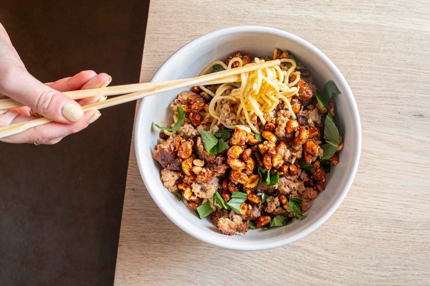 A hand using chopsticks lifts noodles from a white bowl filled with noodles, ground meat, leafy greens, and crunchy toppings, placed on a light wooden table.