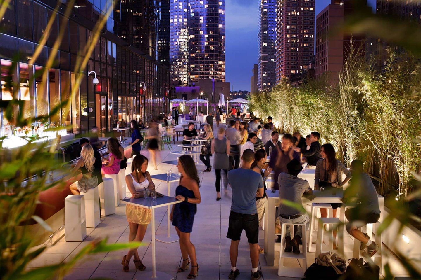 People gather and socialize at a lively rooftop bar in a city at dusk, with lights from tall buildings in the background and greenery lining the terrace. Some guests sit or stand at tables, enjoying drinks and conversation.