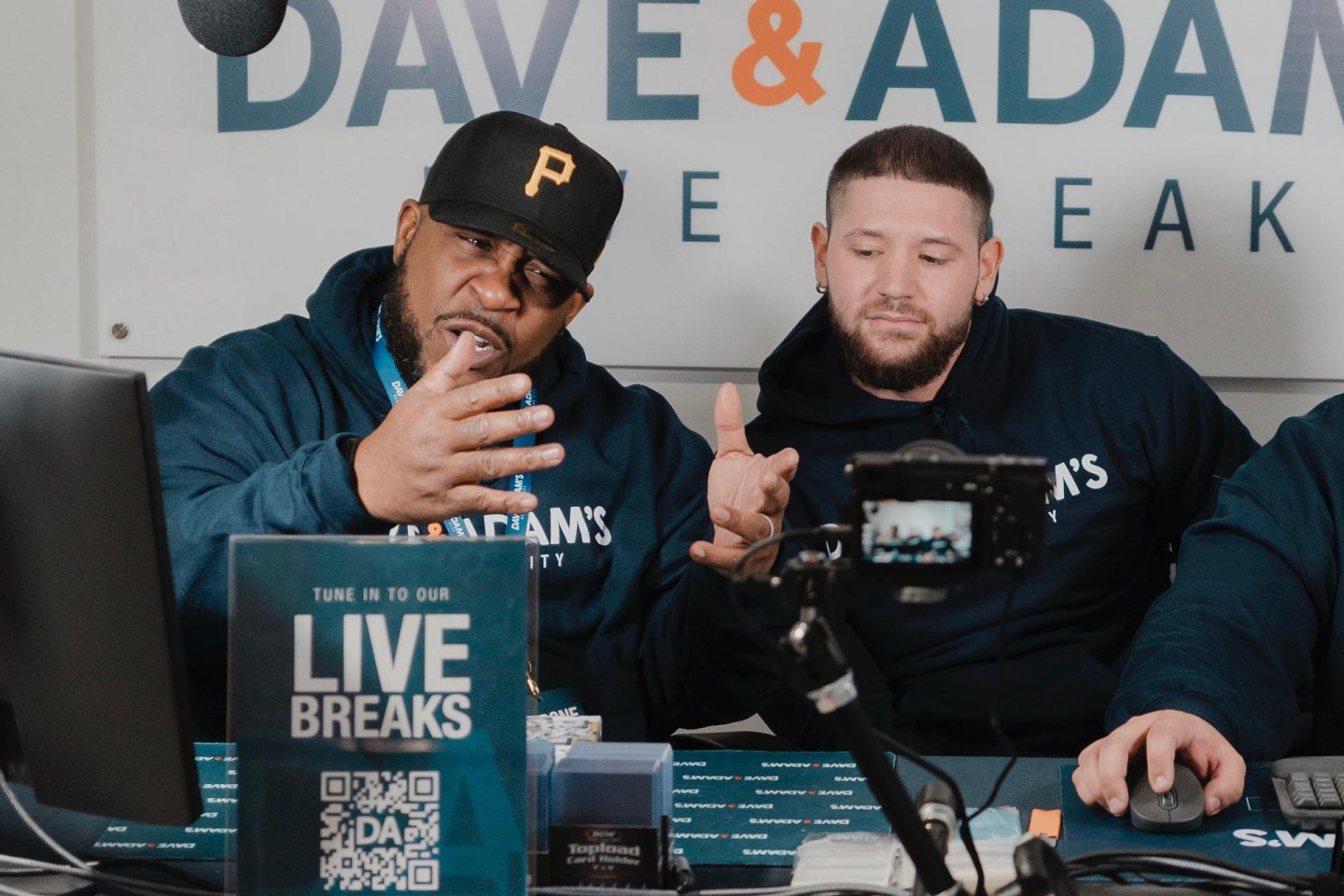 Two men in matching navy hoodies and a cap with a "P" logo sit at a desk with streaming equipment, conducting a live break event. They're engaged, with one gesturing expressively. A sign reads "Tune in to our LIVE BREAKS".