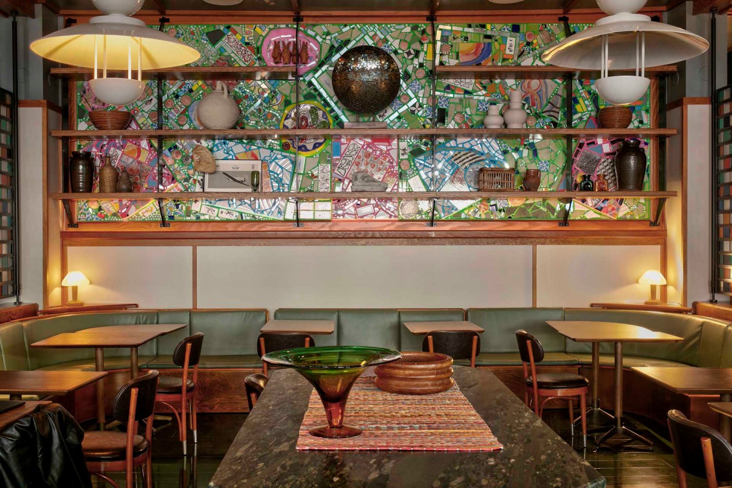 Cozy café with wooden tables and chairs, green cushioned benches, pendant lights, and shelves displaying vases and decor. A colorful, abstract mural decorates the back wall behind the shelves.