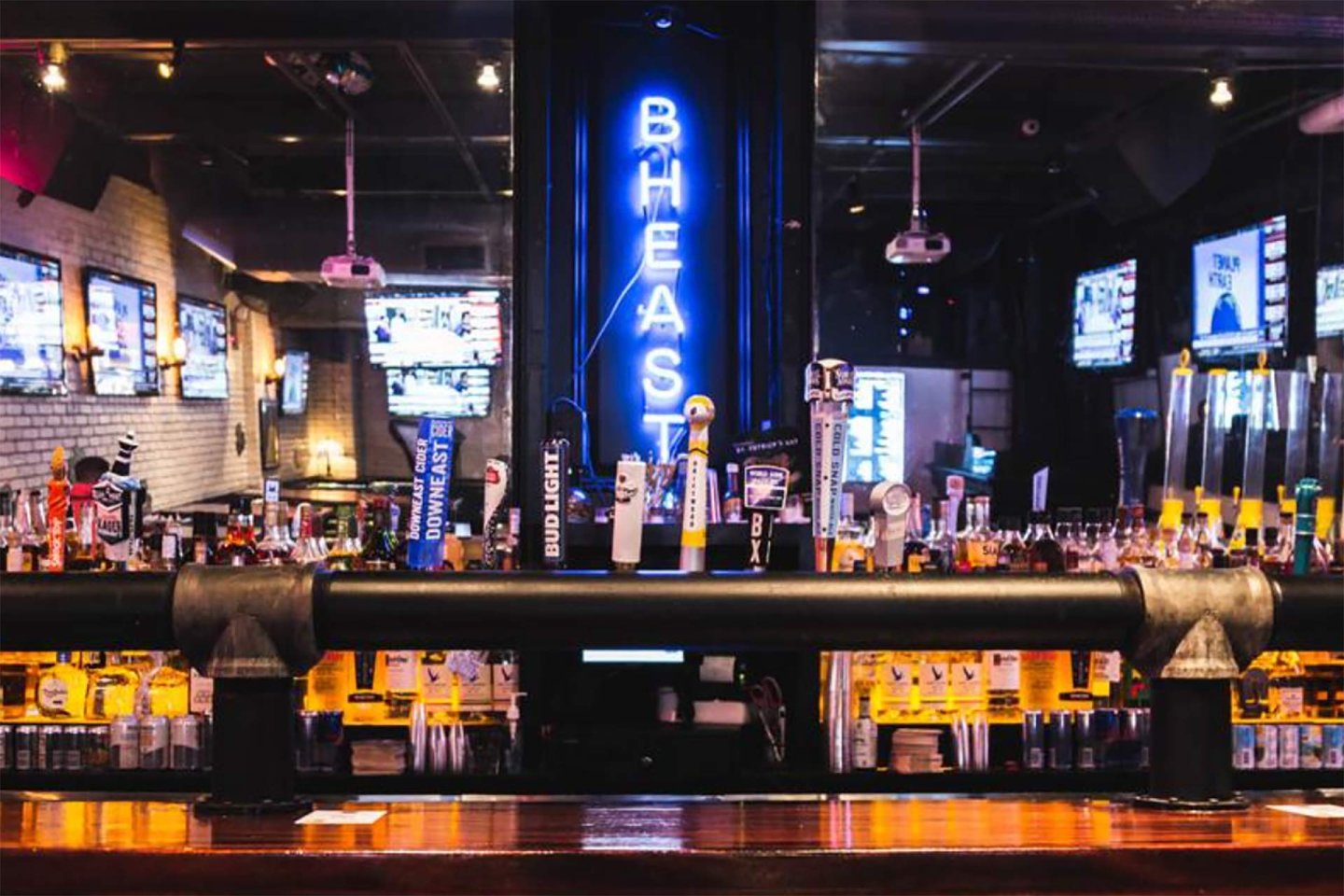 A bar with various beer taps and bottles lined up, illuminated by neon blue "BHEAST" sign in the background. Multiple TV screens display sports channels, creating a lively and modern atmosphere.