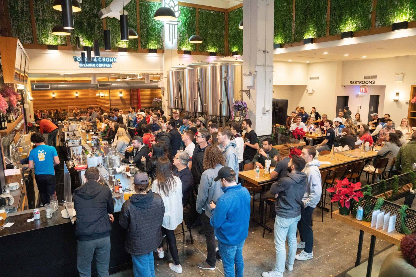 A busy brewery with people gathered around the bar and seated at tables. Large steel tanks are visible in the background. The room is decorated with greenery and bright lights, creating a lively atmosphere.