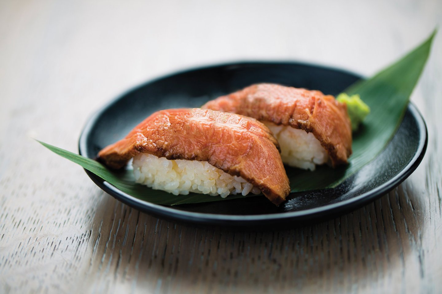 Two pieces of sushi topped with seared meat are placed on a green leaf on a black plate, set on a light wooden surface.