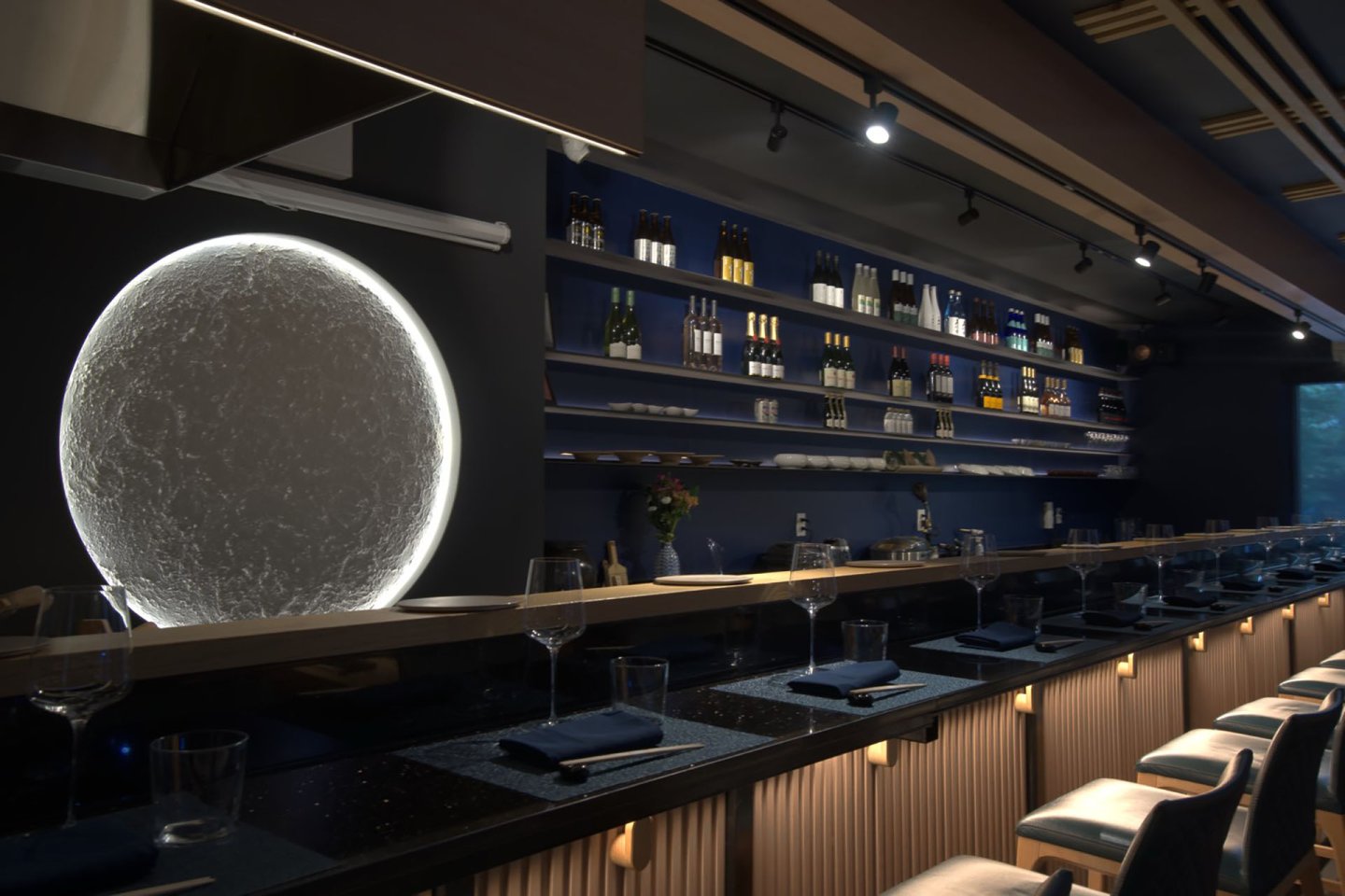 A modern bar with a dark blue interior, illuminated shelves of bottles, a row of stools, set tables, and a large circular light feature resembling the moon on the wall.