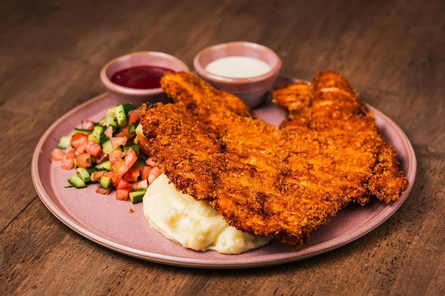 A plate with breaded and fried chicken cutlets on mashed potatoes, a side of diced cucumber and tomato salad, and two small bowls of dipping sauce, on a wooden table.