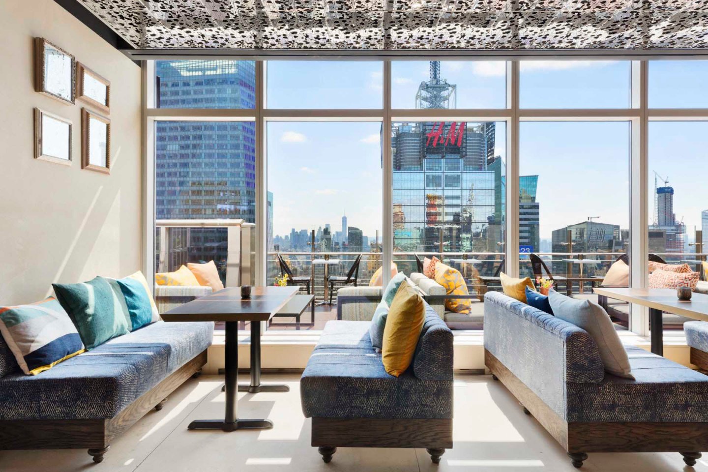 A modern rooftop lounge with blue cushioned seating and colorful pillows. Large windows feature a cityscape view, including tall buildings and a prominent 'H&M' sign. Tables are set for seating, and the ceiling has artistic patterns.