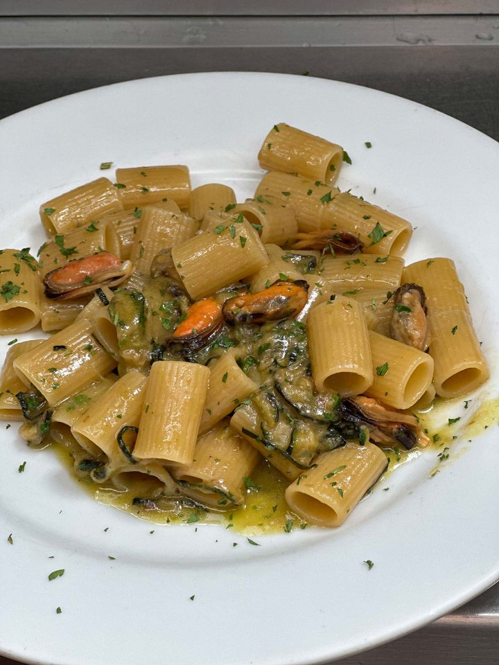 A white plate of rigatoni pasta mixed with mussels and zucchini in a light herb sauce, garnished with chopped parsley.