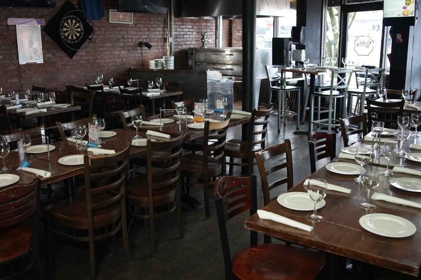 Warm restaurant dining room with dark wood tables set with plates, napkins, and wine glasses, surrounded by wooden chairs, exposed brick walls, a dartboard, and large front windows.