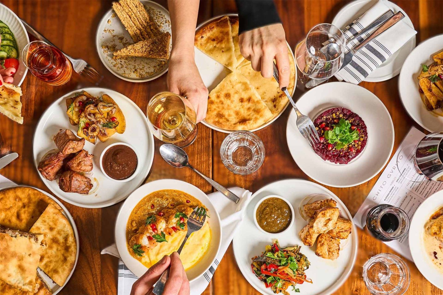 A table filled with various dishes, including bread, grilled meats, dips, and salads, with two people reaching for food and drinks, creating a lively dining atmosphere.