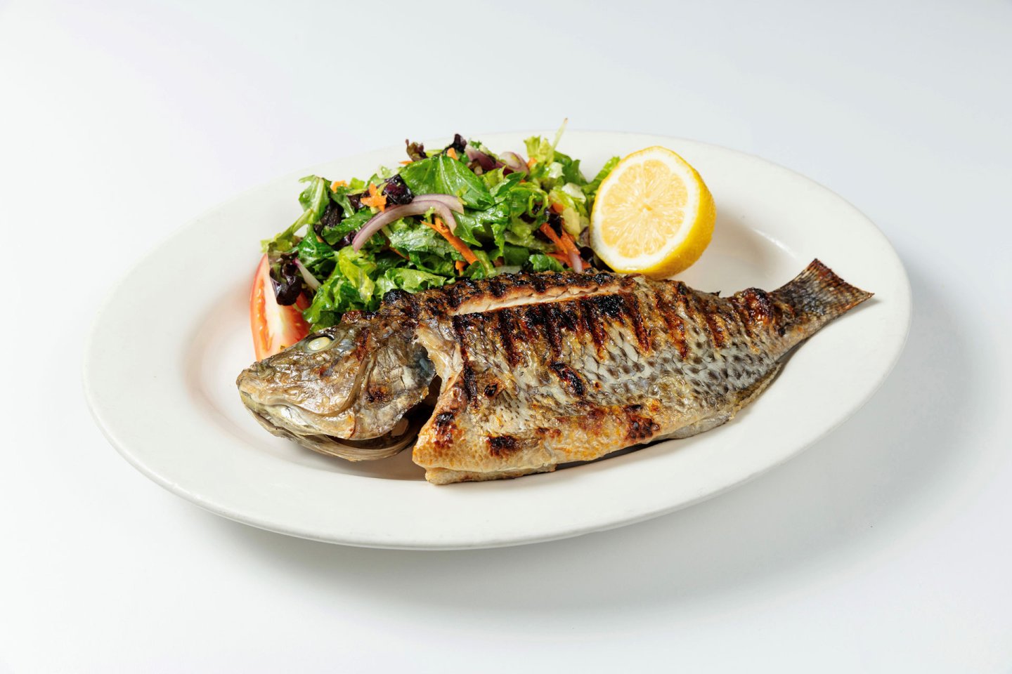 A grilled whole fish is served on a white plate with a fresh green salad and half a lemon on the side, against a plain white background.