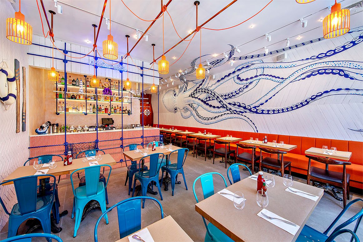A modern restaurant interior featuring teal metal chairs, orange cushioned benches, and hanging woven light fixtures. The walls showcase a blue octopus mural. The bar at the back is stocked with bottles and has a hanging light system with red wires.
