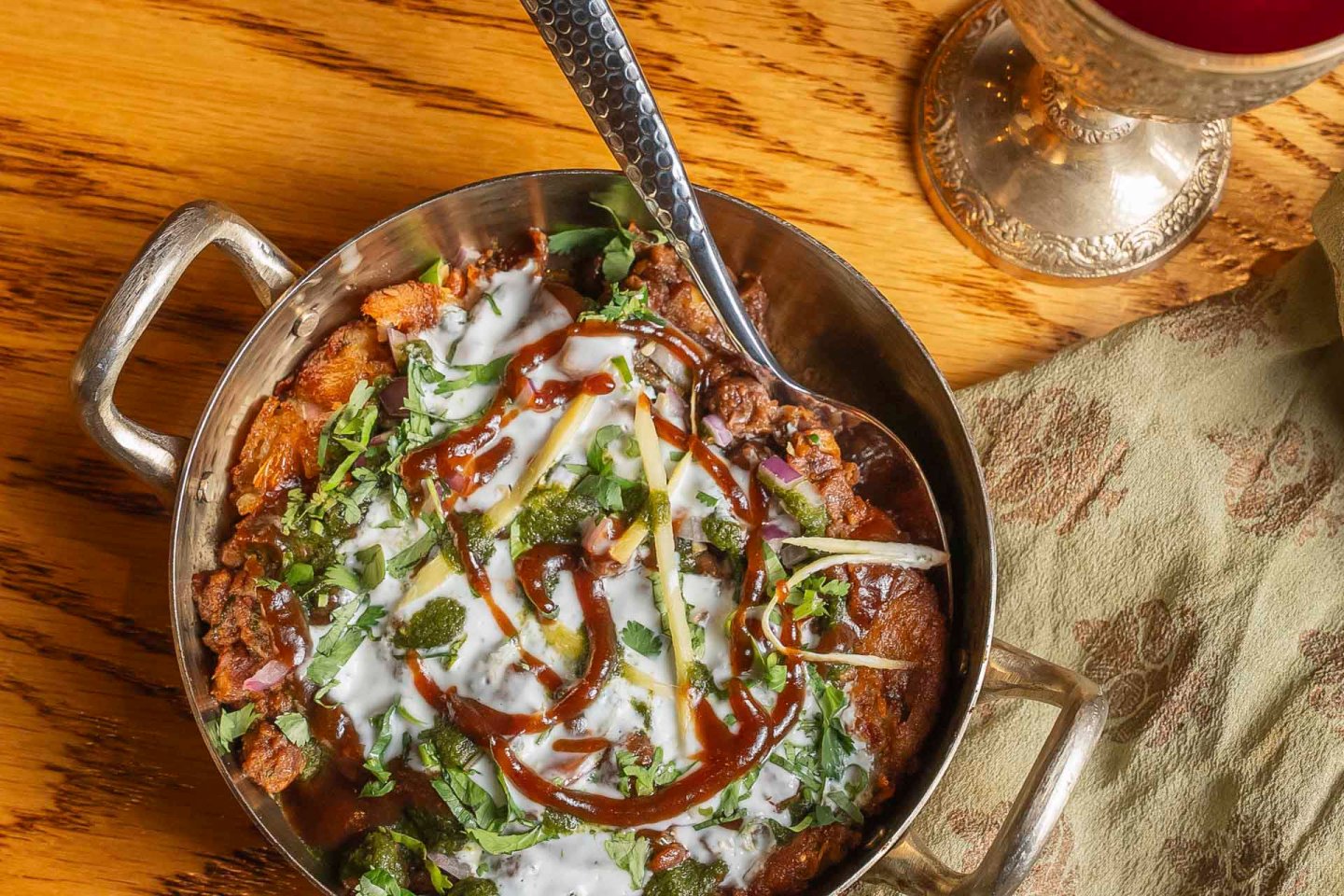 A metal dish filled with chaat, topped with yogurt, tamarind sauce, and chopped herbs, rests on a wooden table. A spoon is placed inside the dish, and a cloth napkin and a decorative cup are next to it.